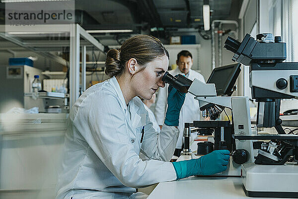 Junge Frau  die einen Objektträger mit menschlichem Gehirn unter dem Mikroskop analysiert  während sie mit Wissenschaftlern im Hintergrund im Labor sitzt