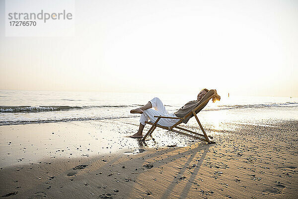 Lächelnde junge Frau  die sich auf einem Klappstuhl am Strand vor einem klaren Himmel bei Sonnenuntergang entspannt
