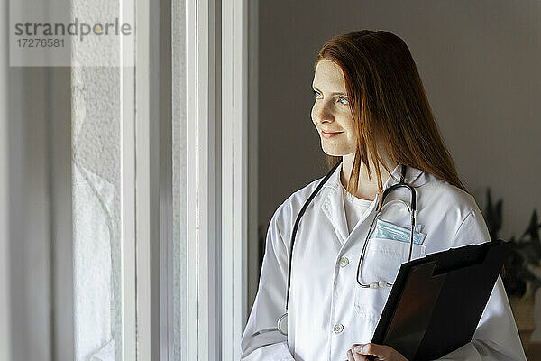 Nachdenkliche junge Ärztin lächelt  während sie durch das Fenster im Büro zu Hause schaut