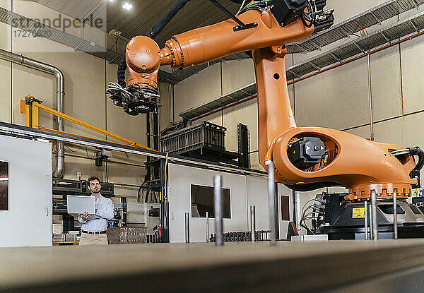 Junger Geschäftsmann mit Laptop  der eine automatische Roboterarm-Maschine in einer Fabrik untersucht