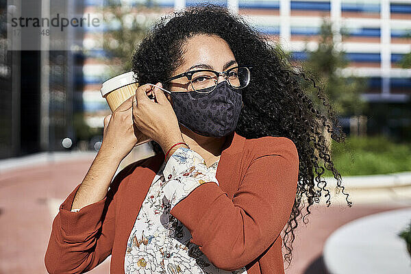 Frau trägt einen Gesichtsschutz und hält einen wiederverwendbaren Bambusbecher in der Stadt an einem sonnigen Tag