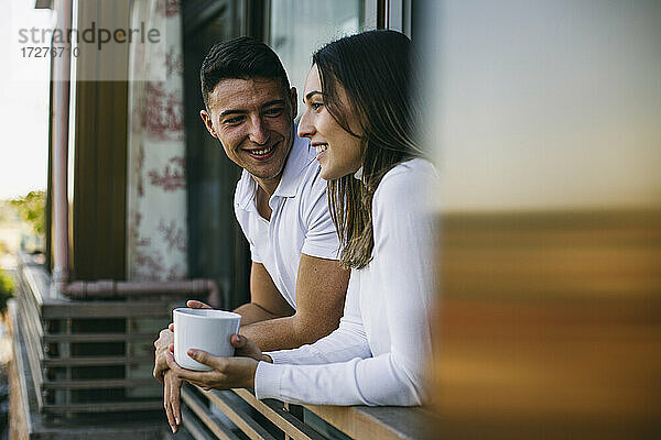 Lächelnder Freund schaut Freundin an  die mit einer Kaffeetasse auf dem Balkon steht