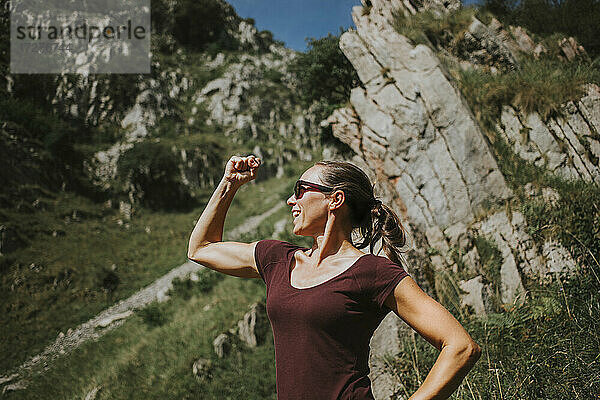 Lächelnde Frau  die Muskeln anspannt  während sie an einem sonnigen Tag vor einer Felsformation steht