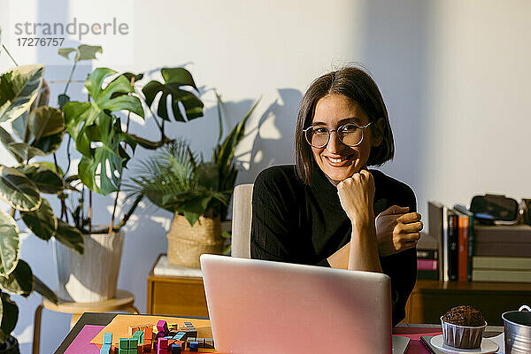 Lächelnde Geschäftsfrau mit Hand am Kinn  die einen Laptop benutzt  während sie zu Hause sitzt
