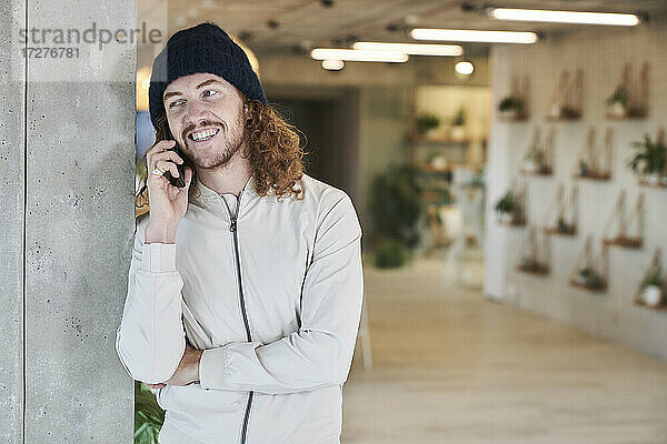 Lächelnder Mann mit Strickmütze  der mit seinem Smartphone telefoniert  während er zu Hause steht