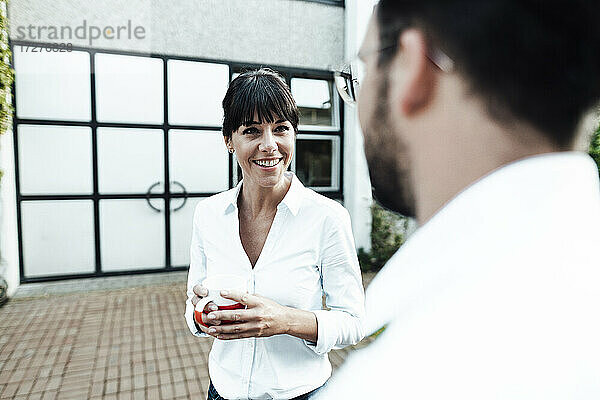 Lächelnde Geschäftsfrau  die eine Kaffeetasse hält und einen männlichen Kollegen während einer Diskussion in der Pause ansieht
