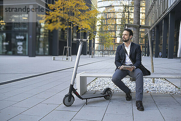Geschäftsmann mit digitalem Tablet sitzt auf einer Bank in der Stadt neben einem E-Scooter