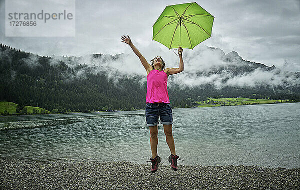 Glückliche reife Frau  die einen Regenschirm hält  während sie in der Regenzeit am Haldenseeufer springt