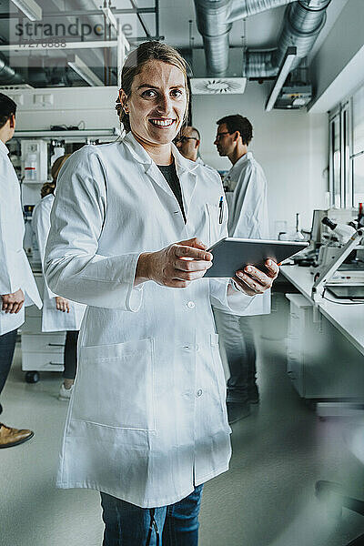 Lächelnde Frau  die ein digitales Tablet benutzt  während sie mit einem Mitarbeiter im Hintergrund in einem Labor steht