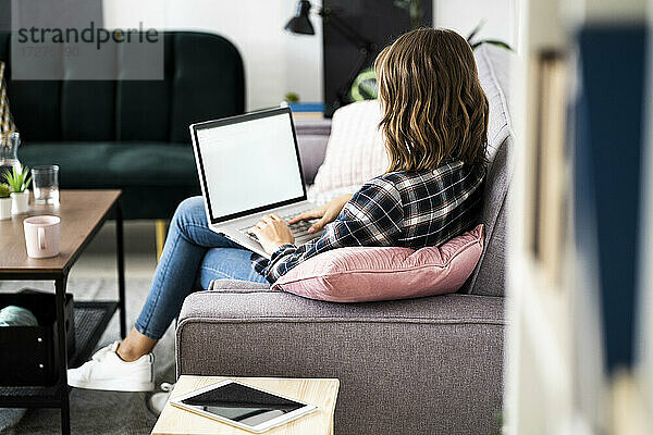 Junge Frau arbeitet am Laptop  während sie zu Hause auf dem Sofa sitzt