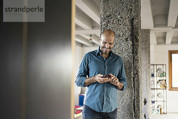 Lächelnder Geschäftsmann  der ein Mobiltelefon benutzt  während er an einer Säule im Büro steht
