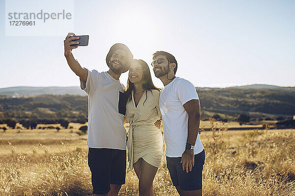 Lächelnde männliche und weibliche Freunde  die ein Selfie mit ihrem Smartphone machen  während sie auf einem Feld im Sommer stehen