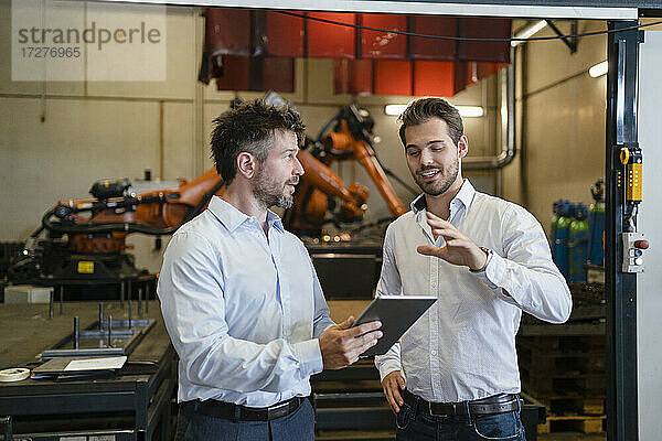 Geschäftsleute mit digitalem Tablet diskutieren  während sie in einer Fabrik vor einer Roboterarm-Maschine stehen
