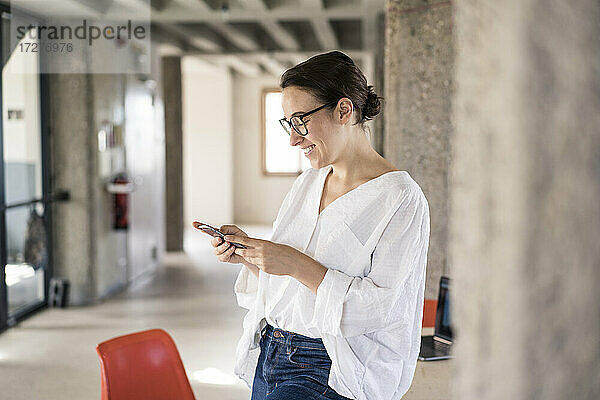 Lächelnde Frau  die ein Mobiltelefon benutzt  während sie im Büro steht