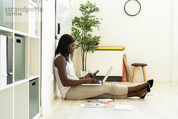 Geschäftsfrau  die Laptop und Mobiltelefon benutzt  während sie im Büro auf dem Boden sitzt