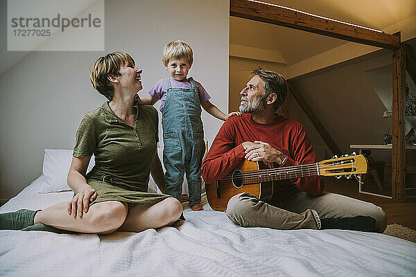 Sohn steht zwischen Mutter und Vater und spielt Gitarre  die zu Hause im Schlafzimmer sitzen