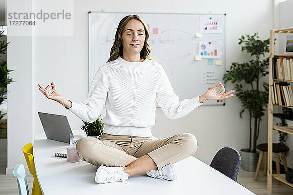 Junge Geschäftsfrau übt Yoga  während sie auf dem Tisch im Büro sitzt