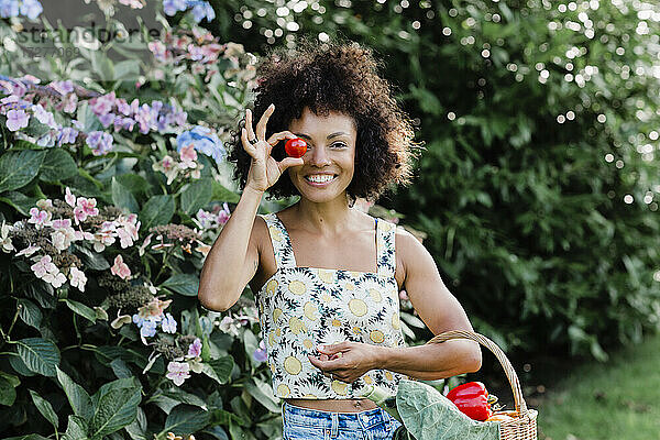 Lächelnde erwachsene Frau hält eine Tomate in der Hand und steht neben einer blühenden Pflanze im Park