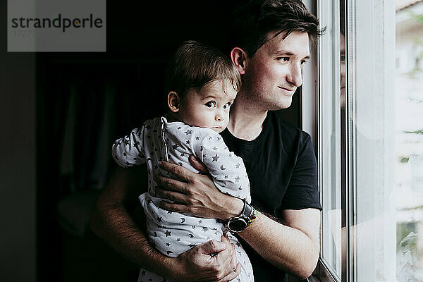 Lächelnder Vater  der seinen Sohn trägt  während er zu Hause durch das Fenster schaut
