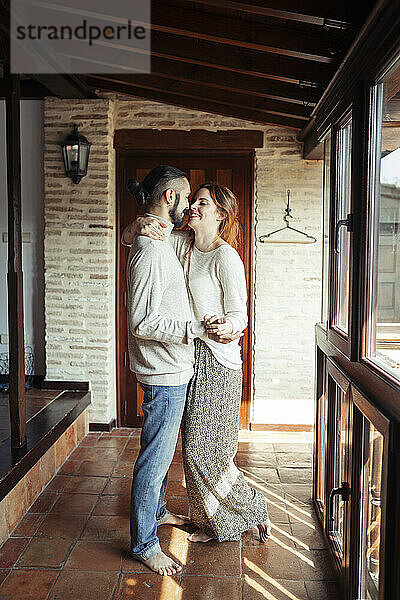 Lächelndes Paar hält sich an den Händen  während es zu Hause am Fenster steht
