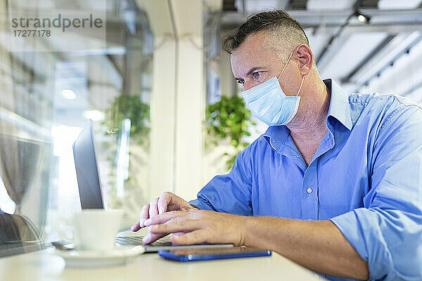 Männlicher Berufstätiger mit Schutzmaske bei der Benutzung eines Laptops in einem Café