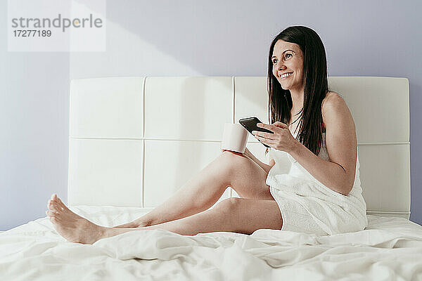 Lächelnde schöne Frau hält Smartphone und Kaffeetasse  während sie auf dem Bett zu Hause sitzt