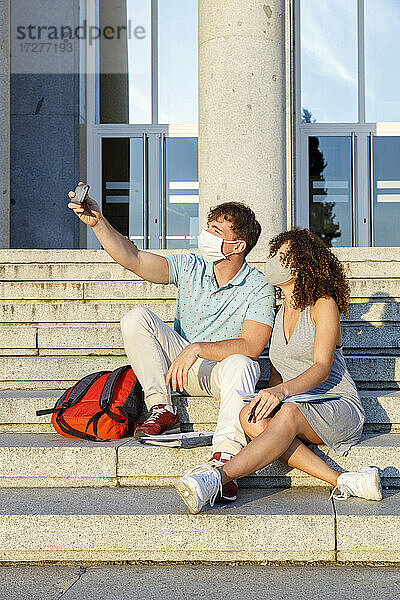 Männlicher Freund macht ein Selfie mit einem Studenten  der eine Schutzmaske trägt  während er auf einer Treppe in der Stadt sitzt