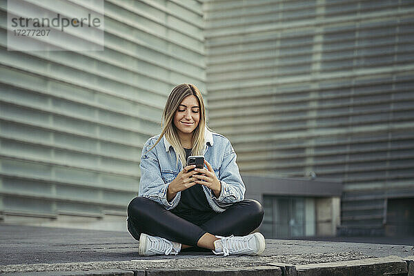Lächelnde junge Frau  die ein Mobiltelefon benutzt  während sie auf dem Gehweg sitzt