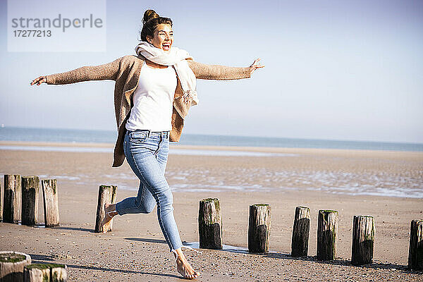 Fröhliche junge Frau mit ausgestreckten Armen  die zwischen Holzpfosten am Strand läuft  während eines sonnigen Tages