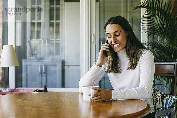 Lächelnde Frau mit Kaffeetasse  die mit ihrem Handy telefoniert  während sie zu Hause sitzt