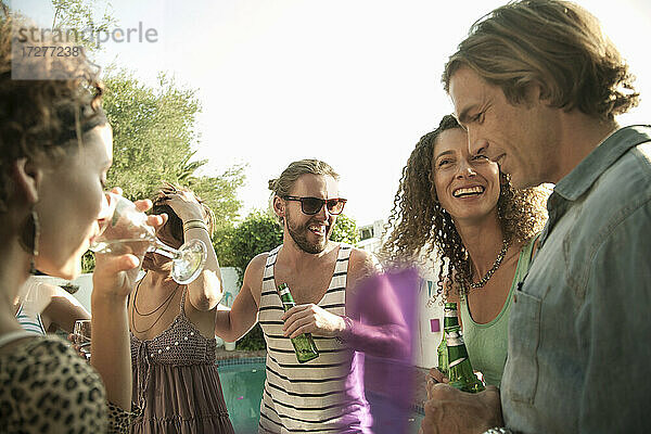 Glückliche männliche und weibliche Freunde genießen den Sommer beim Trinken von Alkohol am Pool gegen den Himmel