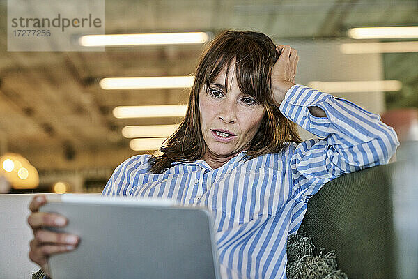 Frau mit schockiertem Gesichtsausdruck  die auf ein digitales Tablet schaut  während sie zu Hause auf dem Sofa sitzt