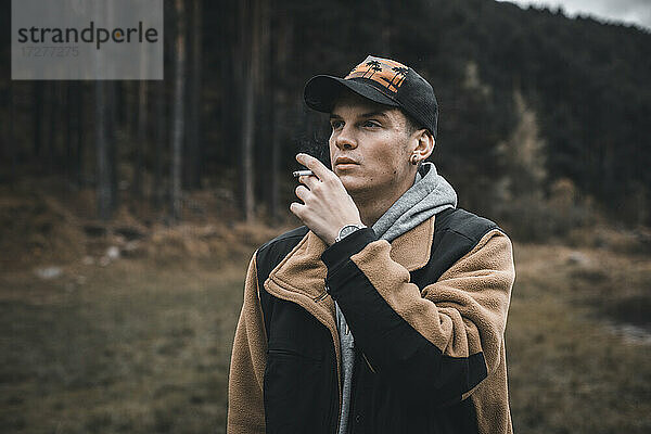 Junger Mann schaut weg  während er rauchend im Wald steht  im Herbst