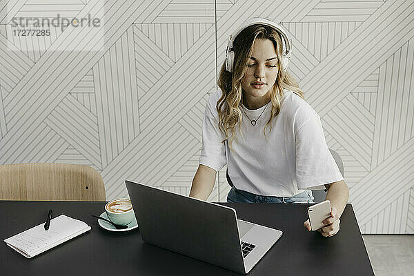 Junge Frau mit Kopfhörern  die an einem Laptop arbeitet und dabei ein Smartphone benutzt  während sie in einem Café sitzt