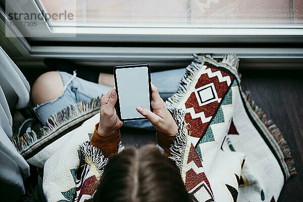 Mädchen mit Decke  das ein Mobiltelefon benutzt  während es zu Hause am Fenster sitzt