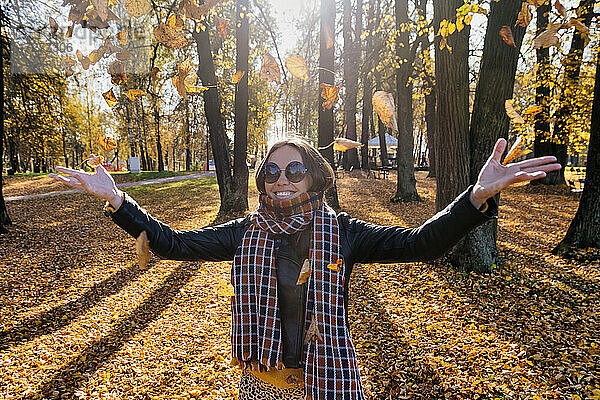 Lächelnde schöne Frau mit ausgestreckten Armen  die trockene Blätter wirft  während sie in einem Park im Herbst steht