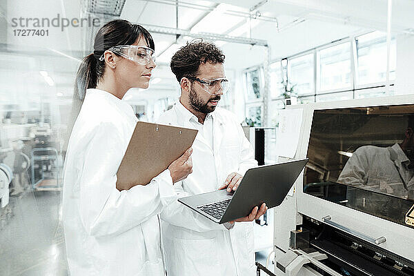 Männliche und weibliche Wissenschaftler mit Laptop und Klemmbrett betrachten Maschinen im Labor