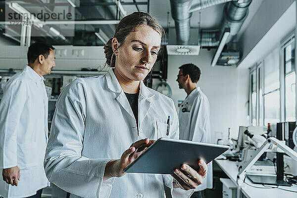 Junge Frau benutzt ein digitales Tablet  während sie mit einem Mitarbeiter im Hintergrund in einem Labor steht