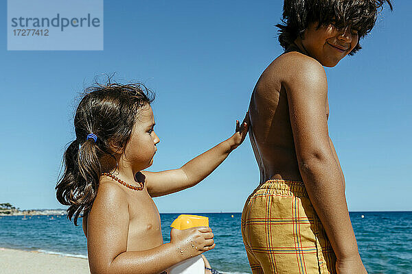 Nettes Mädchen  das Sonnencreme auf den Rücken seines Bruders aufträgt  während es einen sonnigen Tag am Strand genießt