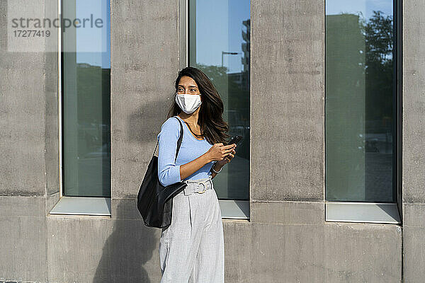 Junge Frau mit Gesichtsschutz  die während COVID-19 an einem Gebäude vorbeigeht