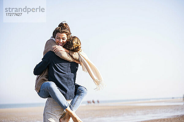 Glückliche junge Frau umarmt Freund am Strand gegen klaren Himmel an einem sonnigen Tag