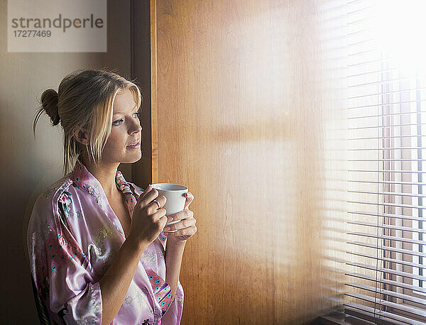 Nachdenkliche junge Frau im Bademantel  die durch das Fenster schaut  während sie zu Hause Kaffee trinkt