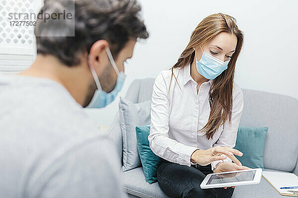 Arzt mit Gesichtsmaske  der ein digitales Tablet benutzt  während er neben einem Mann im Büro sitzt