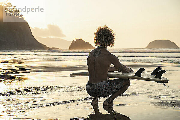 Hemdloser junger Mann mit Surfbrett  der während des Sonnenuntergangs am Meeresufer kauert