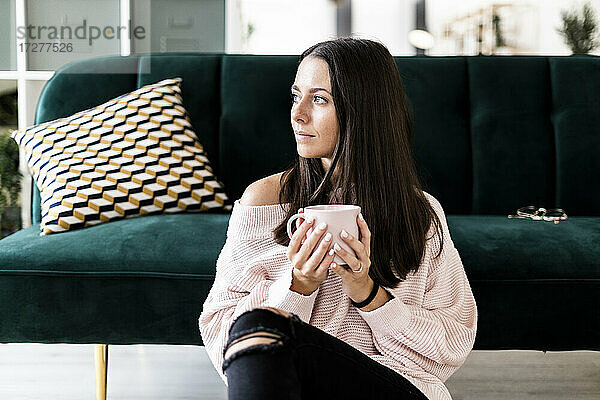 Nachdenkliche junge Frau  die wegschaut  während sie mit einer Kaffeetasse auf dem Sofa zu Hause sitzt