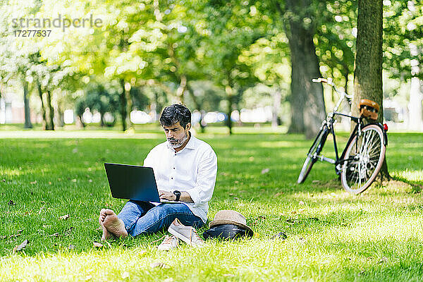 Älterer Mann  der einen Laptop benutzt  während er im öffentlichen Park im Gras sitzt