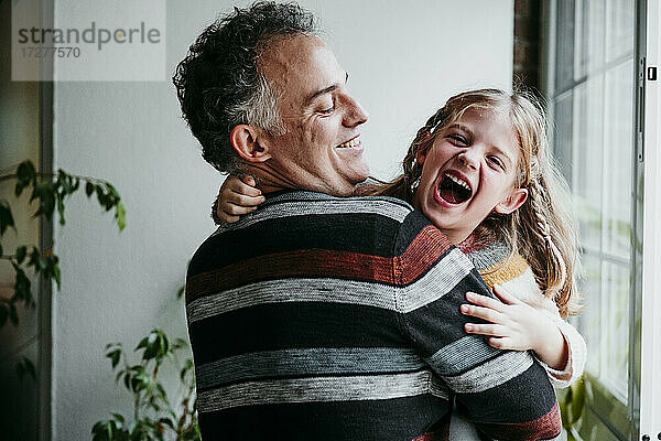 Lächelnder Vater mit verspielter Tochter am Fenster stehend zu Hause