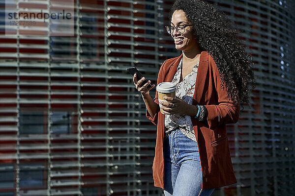 Junge Frau  die lächelnd ein Smartphone benutzt und einen wiederverwendbaren Becher hält  während sie gegen ein Gebäude läuft