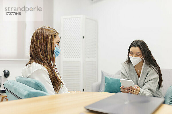 Patient mit Gesichtsmaske  der ein digitales Tablet benutzt  während er bei einem Psychologen im Büro sitzt