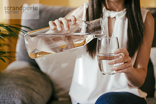 Frau gießt Wasser aus einem Glas in ein Trinkglas  während sie zu Hause sitzt